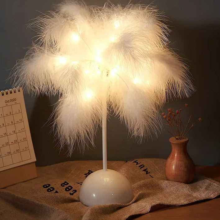 Stilvolle 'LuceSoffusa' Nachttischlampe mit warm leuchtenden, weißen Federn auf einem bronzefarbenen Standfuß, neben einem Kalender, betont durch eine Fernbedienung im Vordergrund.